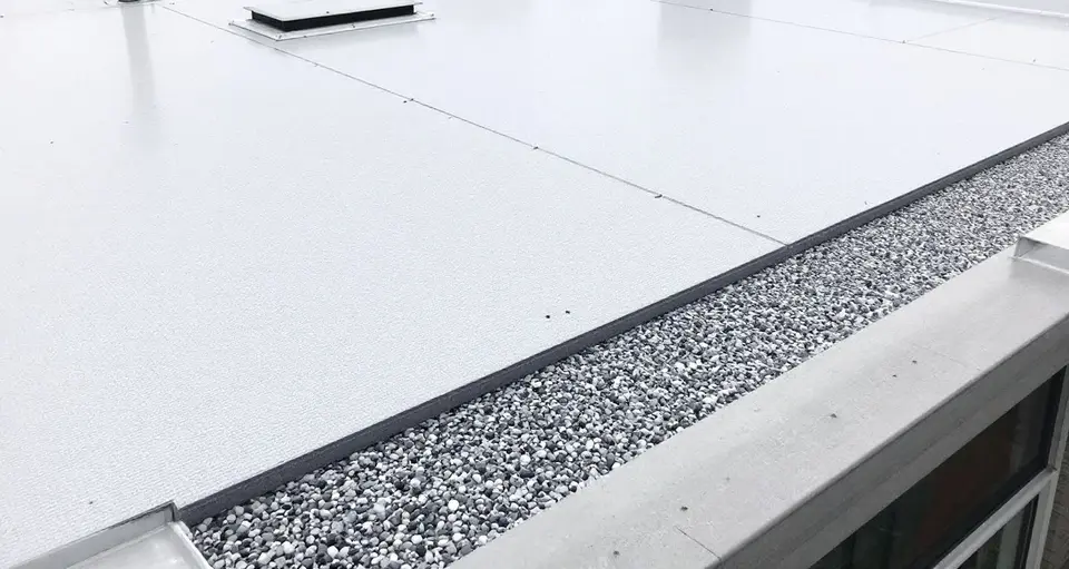Groot plat dak van een appartementengebouw met lichtgrijze PVC dakbedekking, een strook grind langs de rand en een dakluik